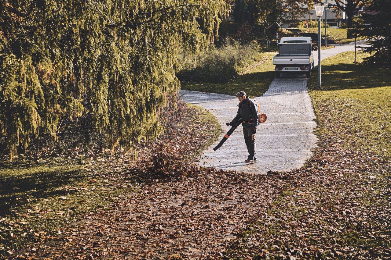 A man in work equipment using a cordless leaf blower to remove leaves from a park path