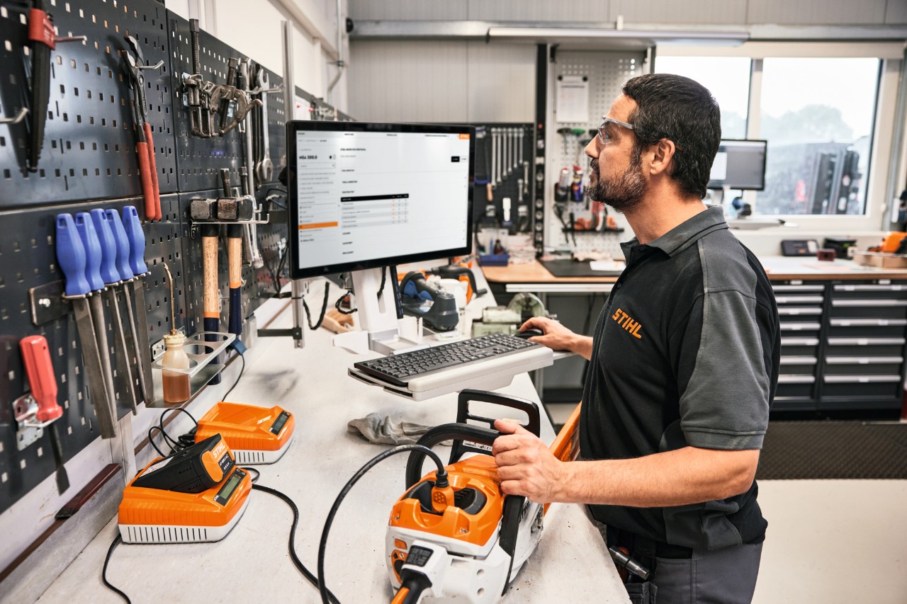 In a workshop, a man stands in the foreground operating a PC. On the table in front of him is a chainsaw