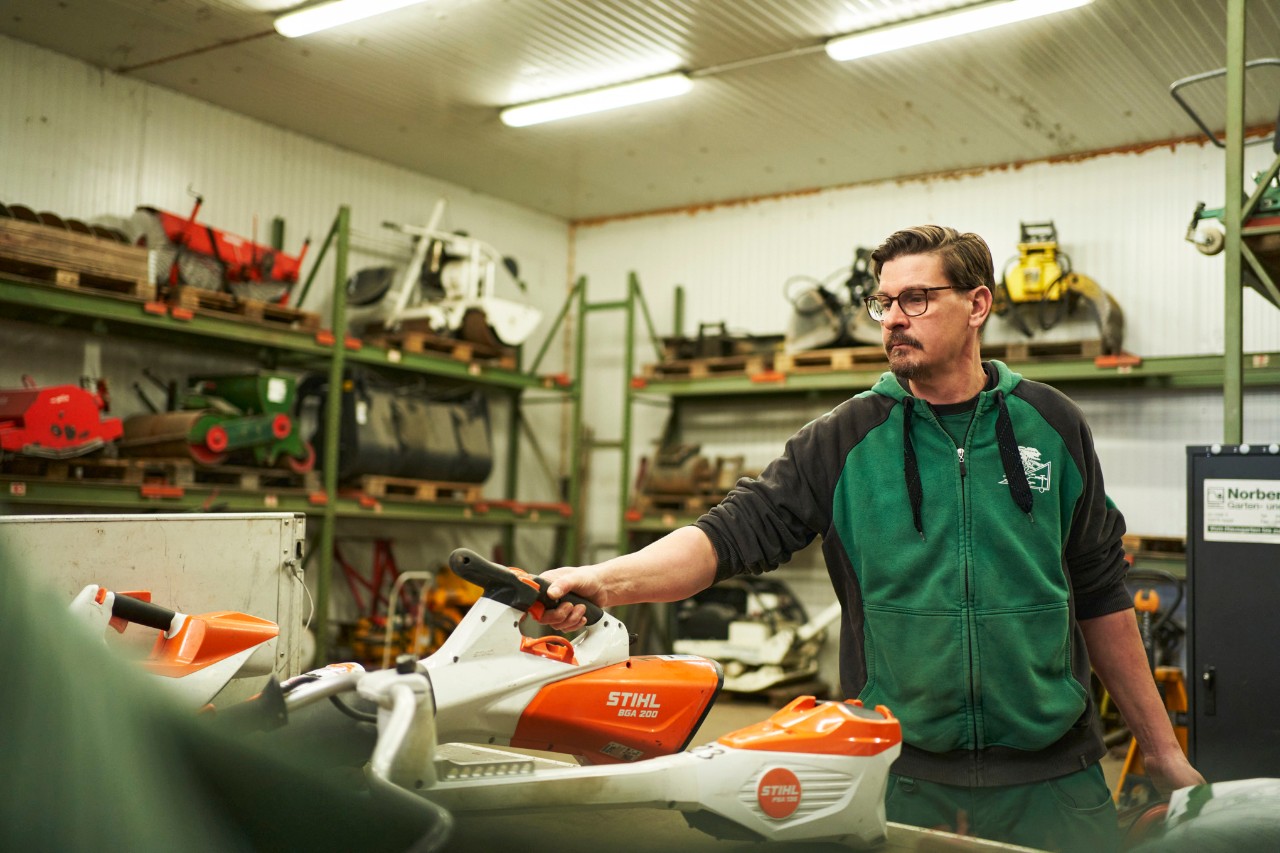 A man in a workshop stands behind a work surface containing STIHL cordless power tools, and reaches for a STIHL BGA 200 cordless leaf blower