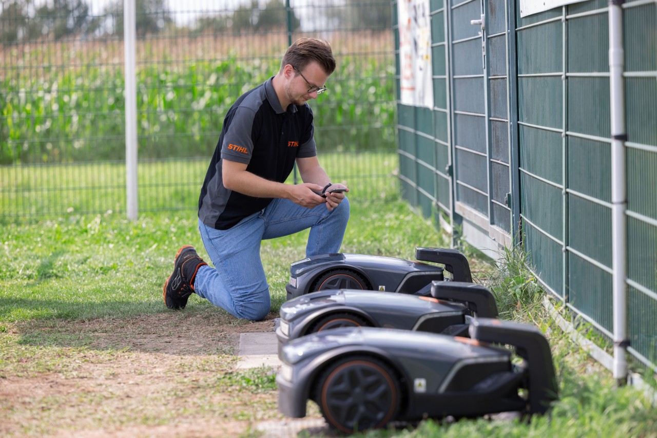 STIHL iMOW® Product Manager Thilo Ehrentreich kneeling next to three robotic mowers with a mobile phone in his hand