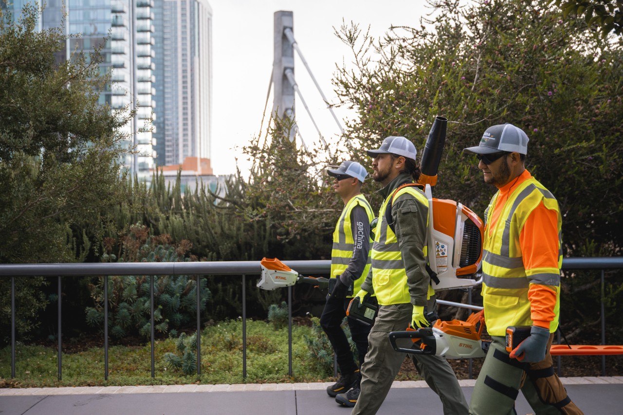 Three landscaping workers walk through an urban city wearing work clothes and holding STIHL power tools.