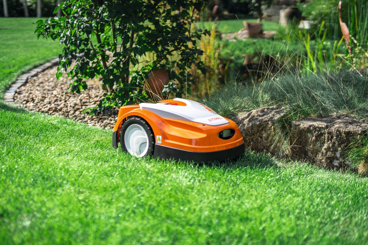 A STIHL iMOW RMI 422 mowing a grassy area in a garden, at the edge of a small stone wall.
