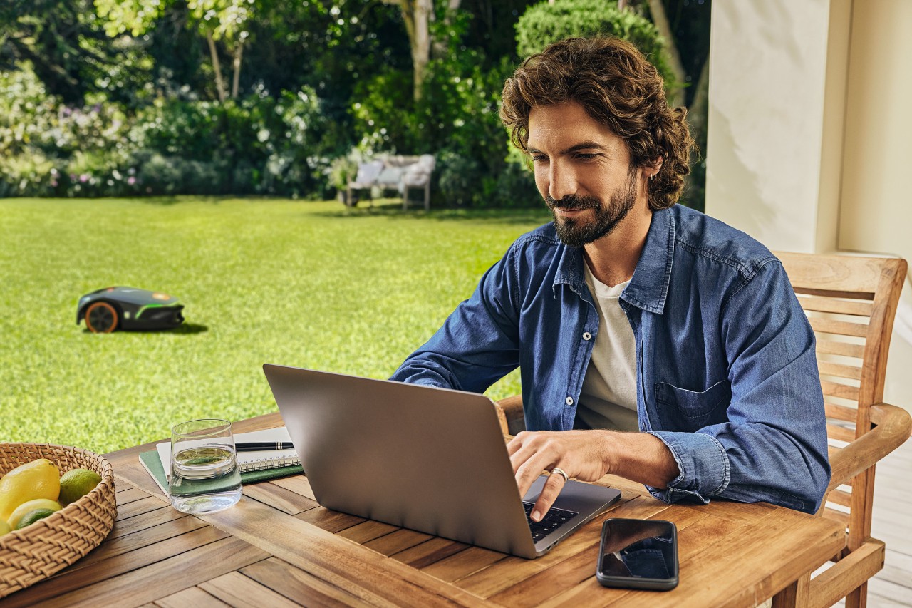 A man sitting on his terrace in front of a laptop and a smartphone, while an iMOW EVO automatically mows the garden in the background.