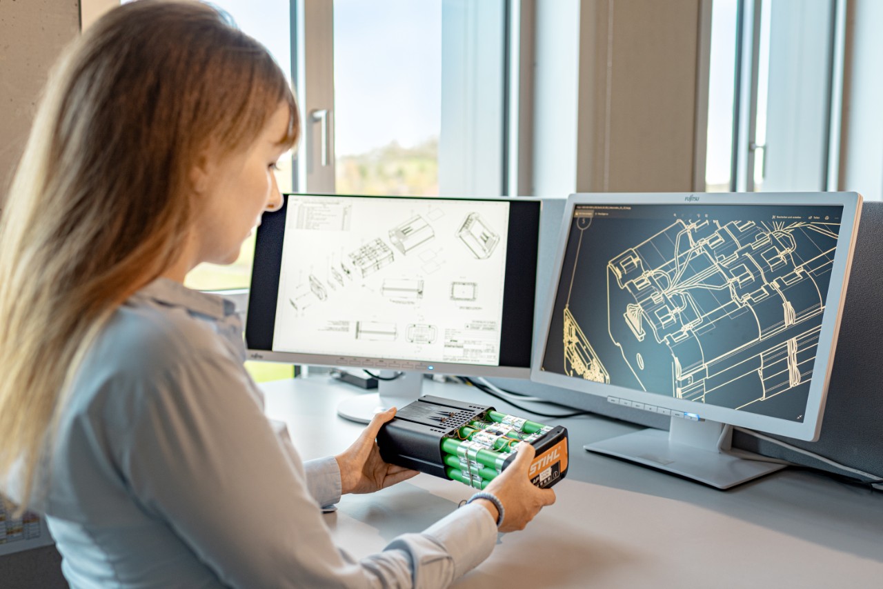 Woman sitting at a desk in front of two miniature monitors with a STIHL battery in her hand